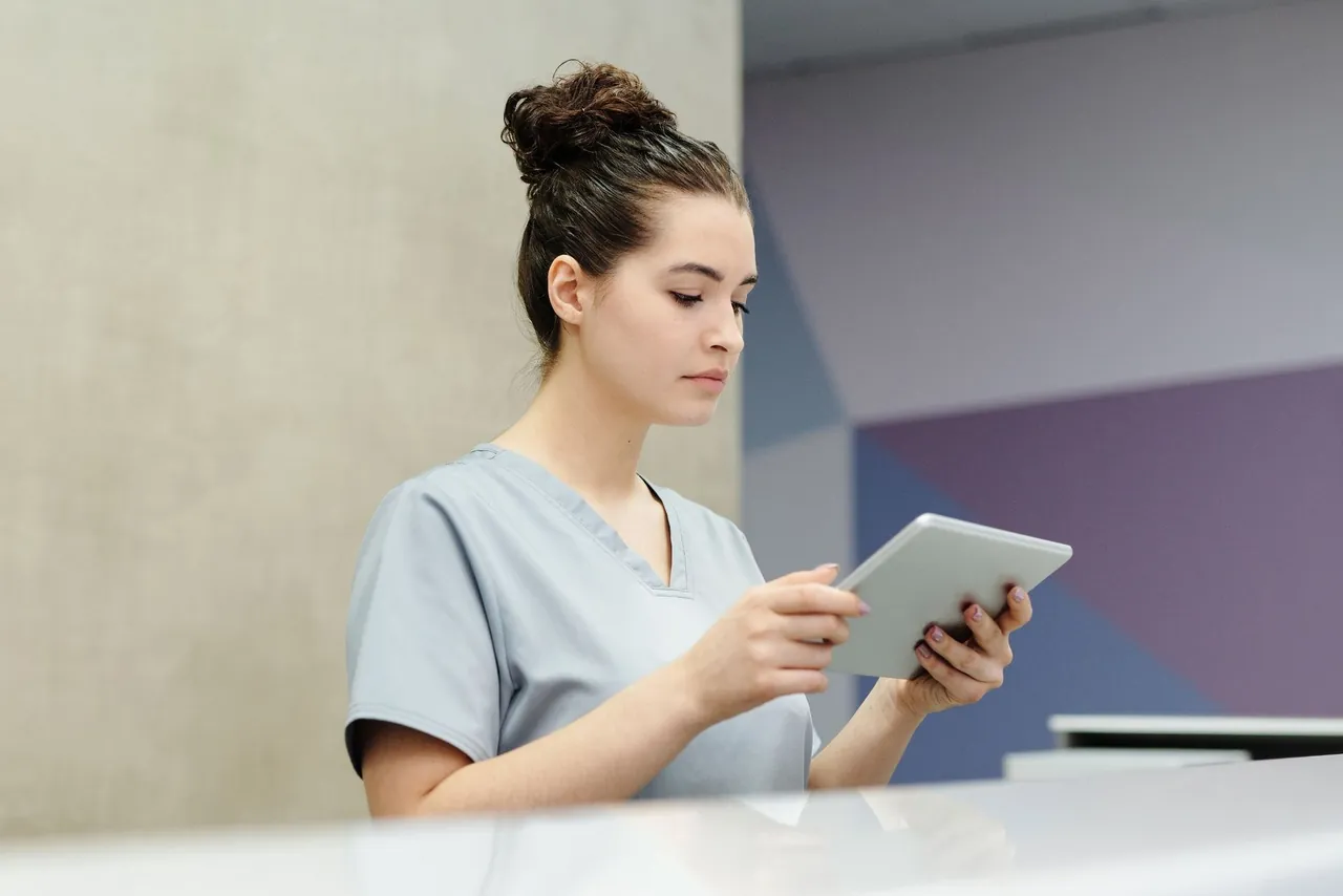 A clinic staffer in scrubs reads a tablet at a front desk in daylight.