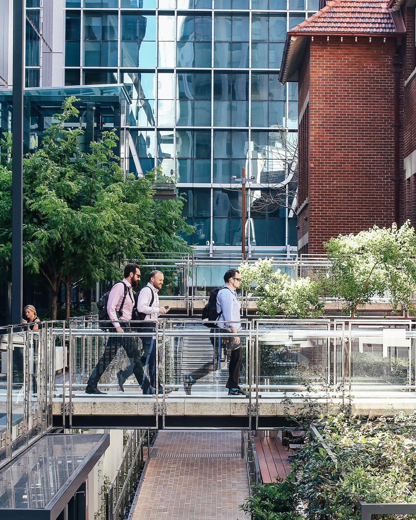 Office workers in business shirts cross a glass footbridge between a glass office tower and a red brick building.