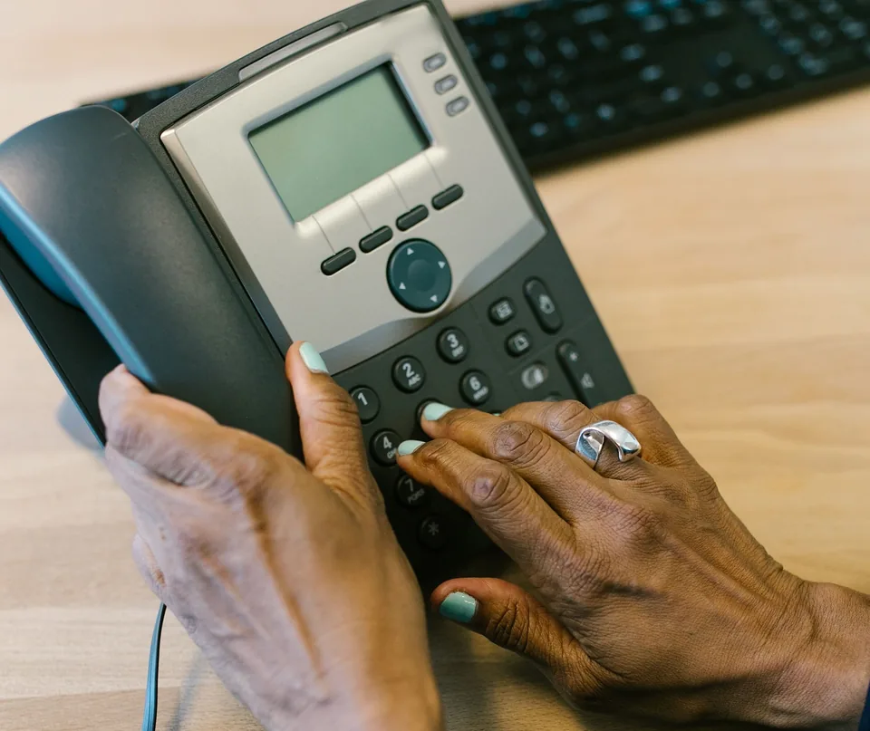 Hands dialing an office desk phone, with a keyboard on the desk behind it.