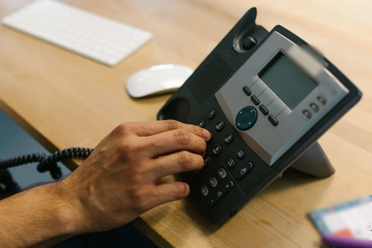 A hand dials a desk phone on a light wood office desk, with a keyboard and mouse behind it.