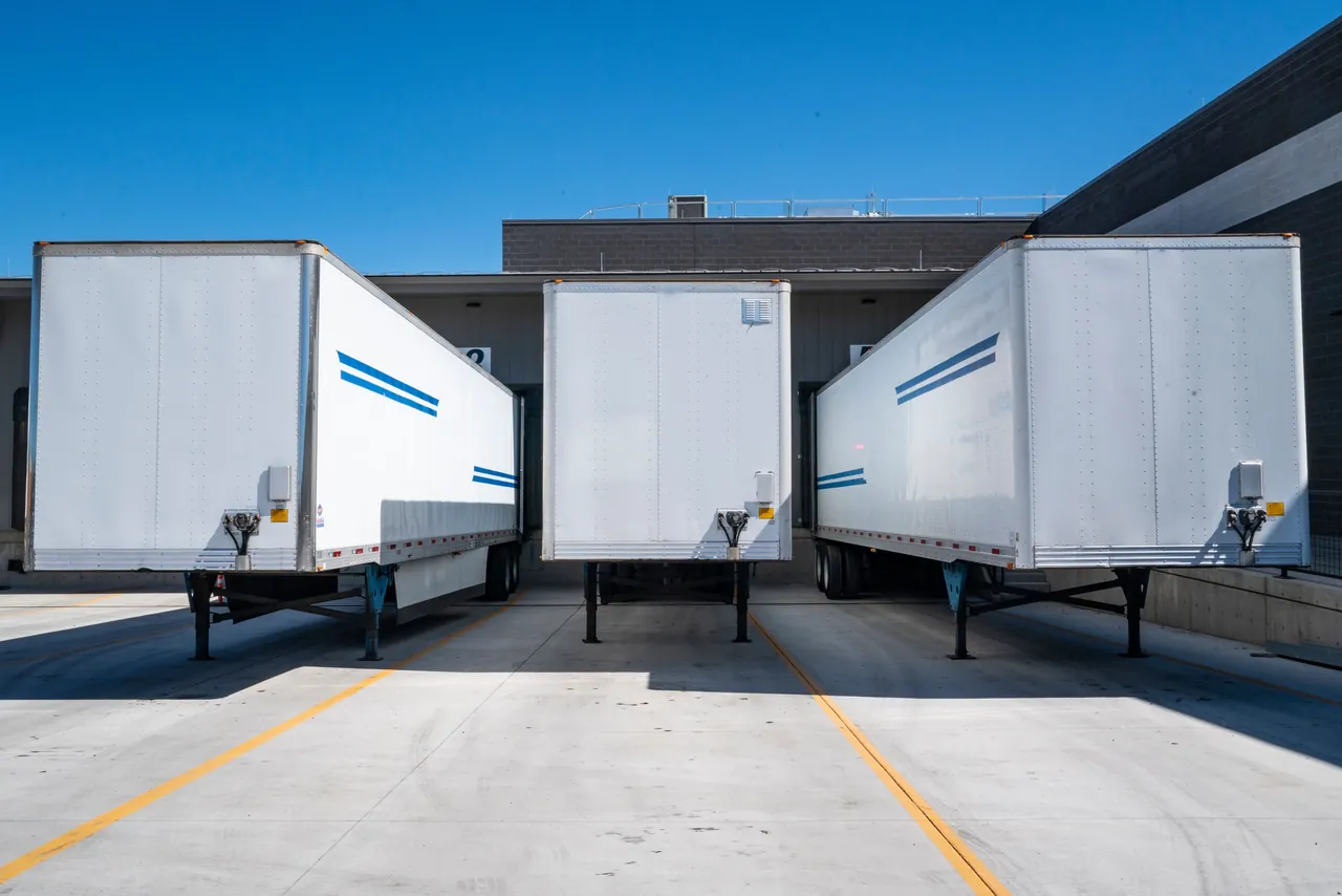Three white semi-trailers backed into the loading docks of a distribution center on a sunny day.