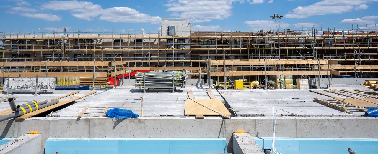 A poured concrete deck with stacked shoring props, timber and conduit on a large building site, a scaffolded building behind it under a blue sky.
