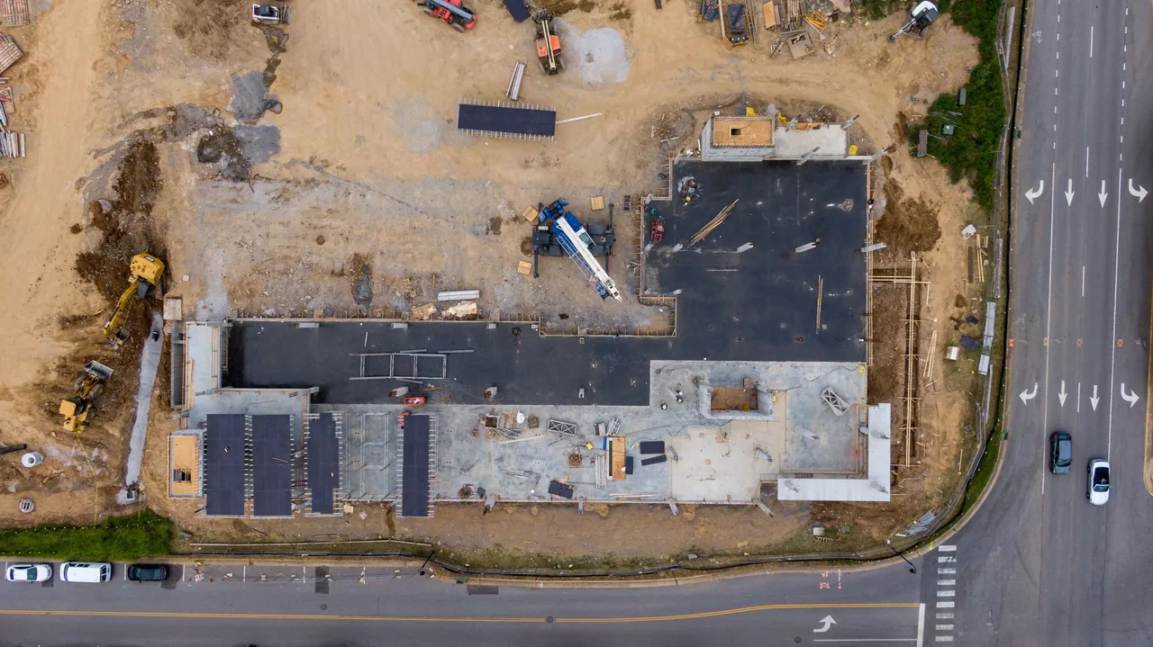 A large building going up beside a road intersection, seen from directly above, with excavators, a mobile crane and stacked materials around the slab.