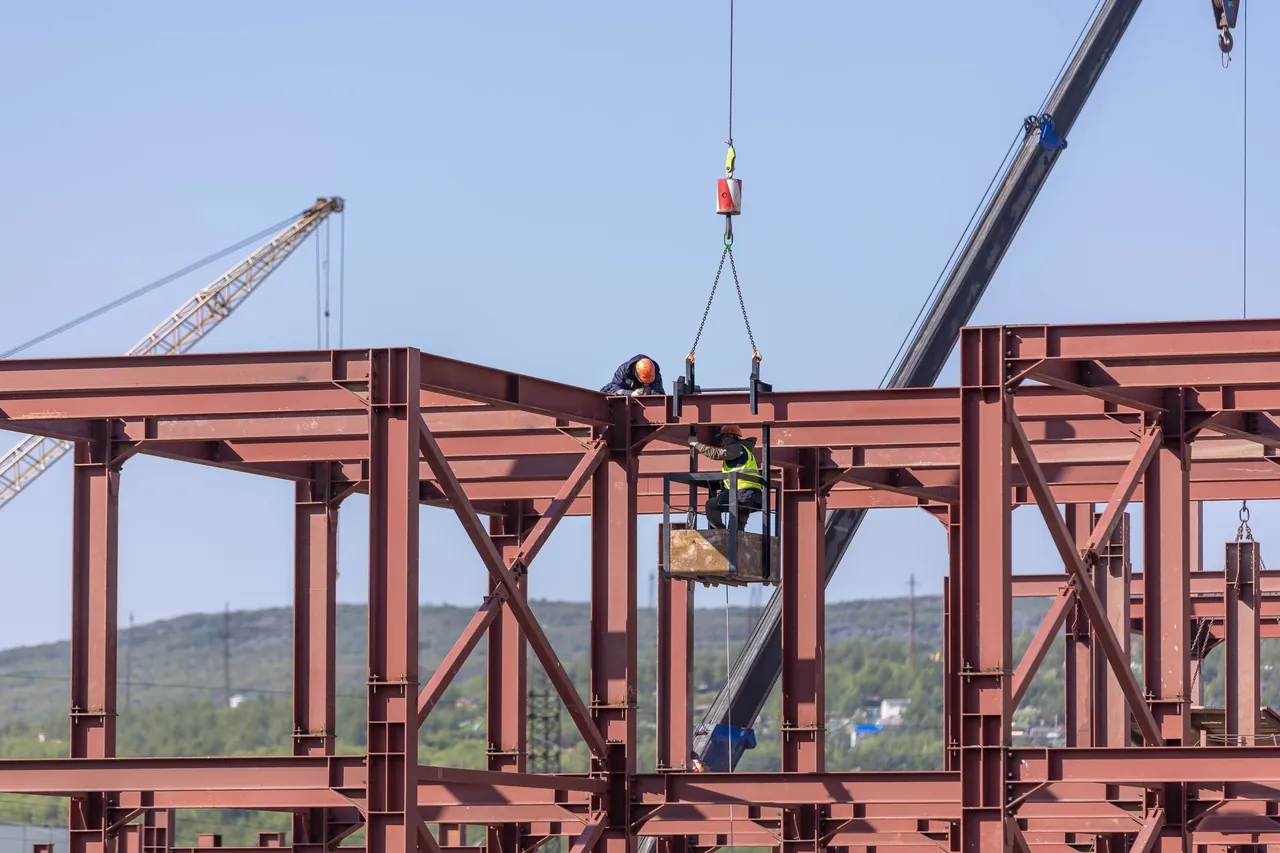Ironworkers bolting the red steel frame of a large commercial building, one on a beam and one in a lift basket, with a crane boom behind them under a clear sky.