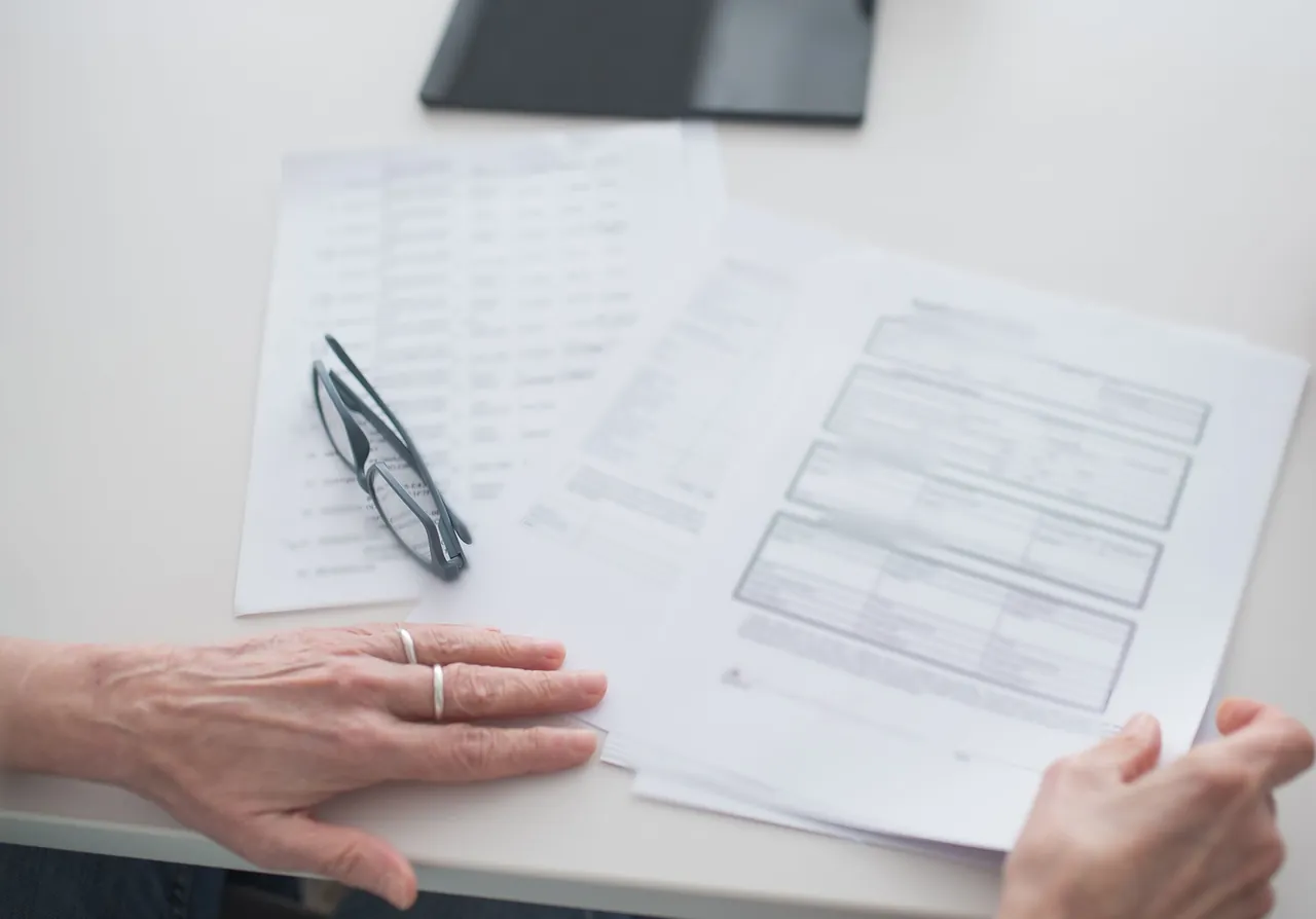 Hands around printed tables and forms at an office meeting table, with reading glasses resting on the pages.
