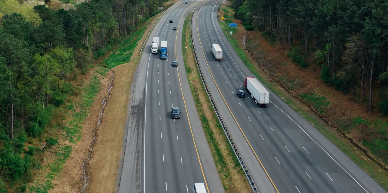 A four-lane interstate through woodland seen from the air, with several tractor-trailers traveling in both directions.