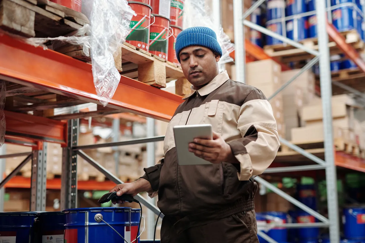 A warehouse worker in a blue beanie checks a company tablet in a pallet-rack aisle, a corded barcode scanner in his other hand.