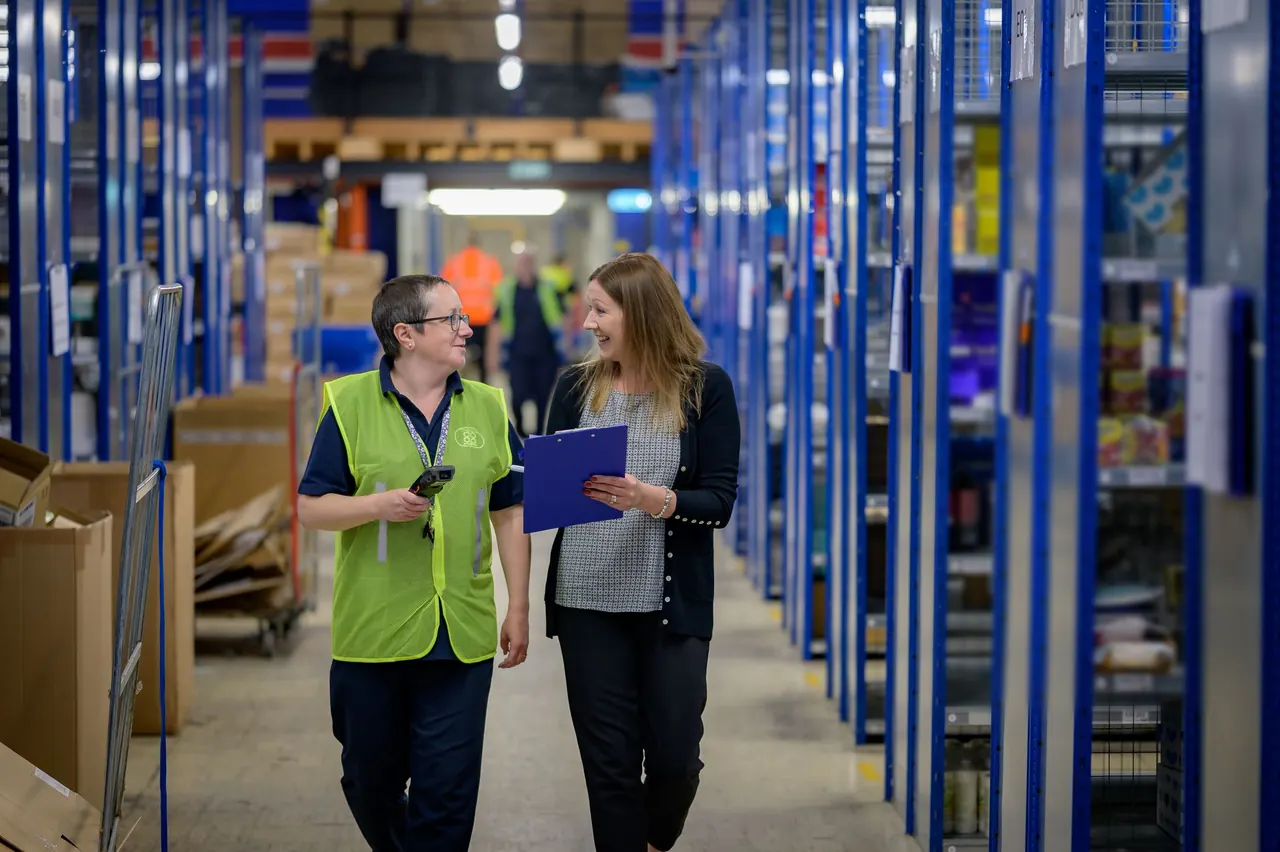 Two staff walk a distribution center aisle between tall blue shelving, one in a high-visibility vest holding a handheld scanner, the other with a clipboard.