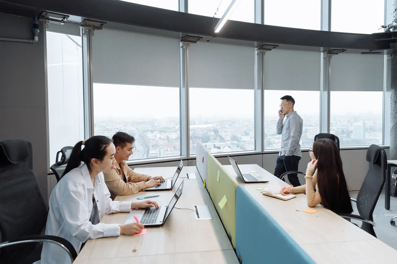 A team works at bench desks with laptops on a high office floor while a colleague takes a call on a company phone by the window, the city below in daylight.