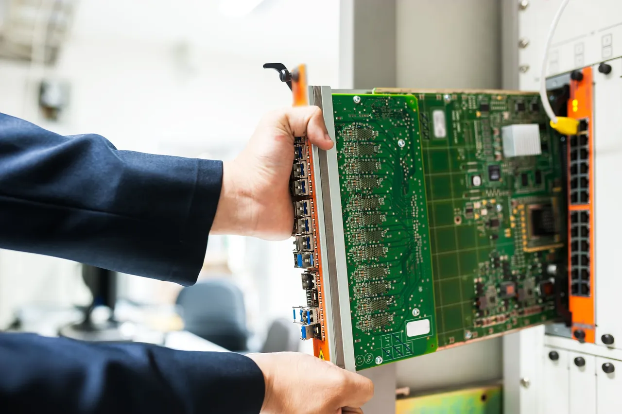 A technician's hands slide a line card into a network equipment shelf in a bright equipment room.