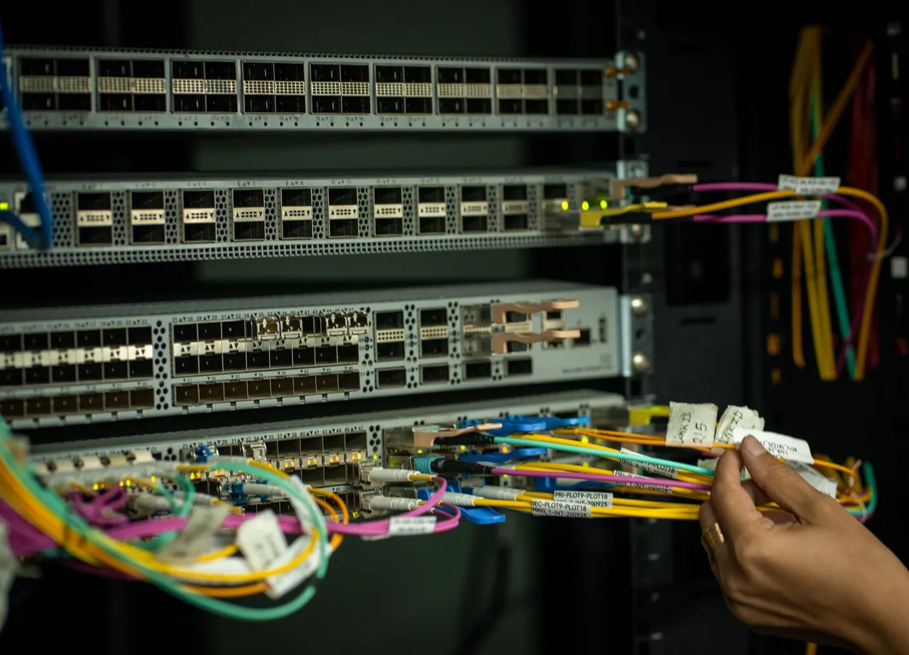 A hand sorts labeled fiber jumpers at a row of network switches in a dark equipment room.