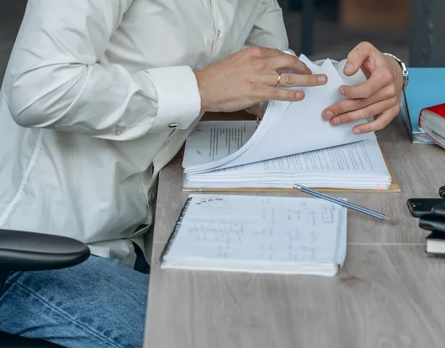 Hands page through a thick stack of printed statement pages on an office desk, beside a notebook and a pencil.