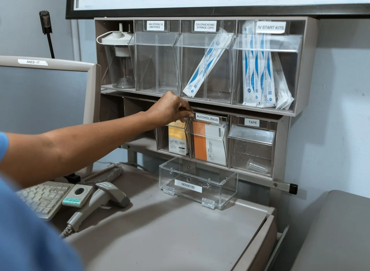 A staff member's hand reaches into labeled supply bins at a clinic workstation, with a monitor and a handset scanner on the counter.