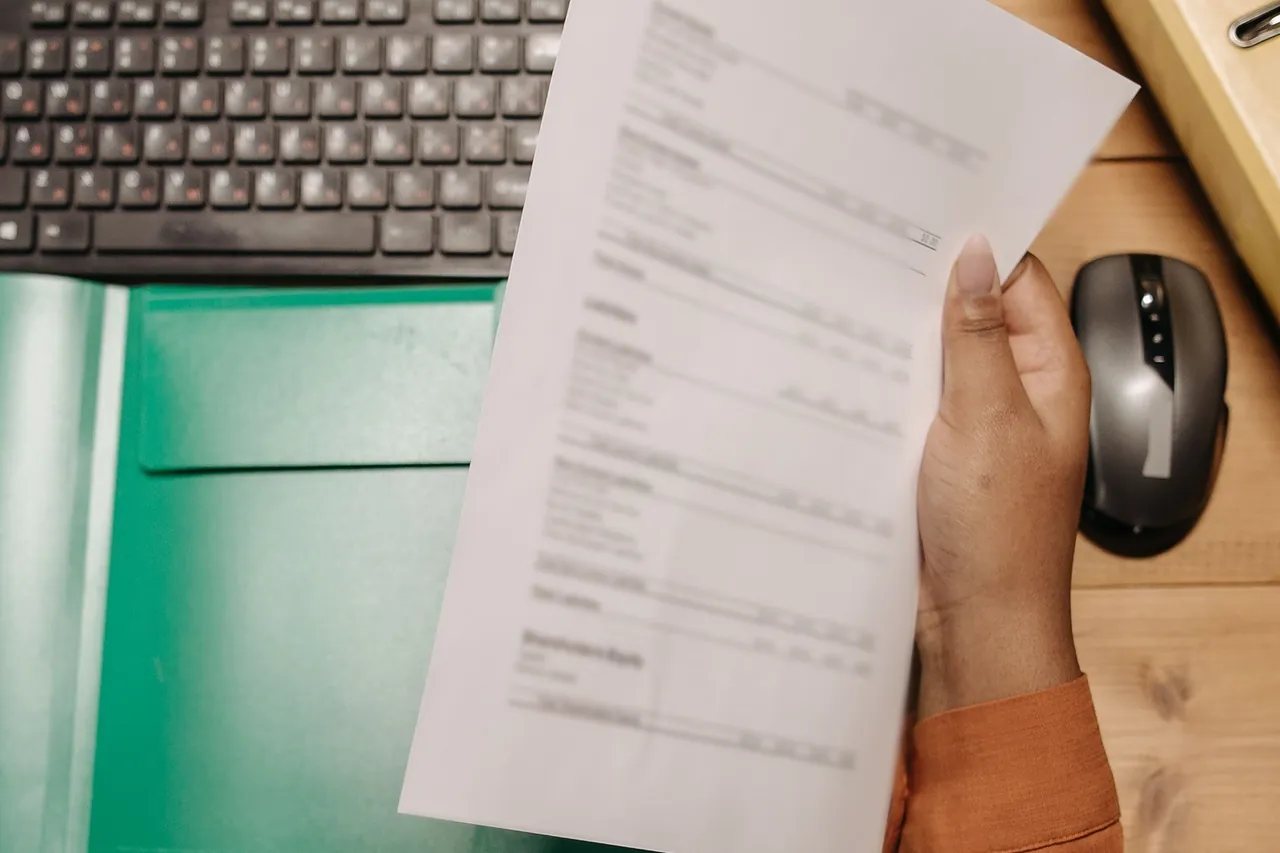 A hand holds a printed statement page with columns over an office desk, beside a keyboard and a green folder.