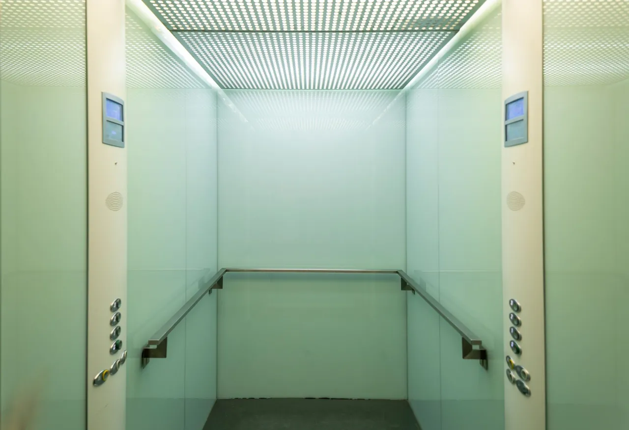 An empty elevator cab with steel handrails and call panels on both sides, in even light.