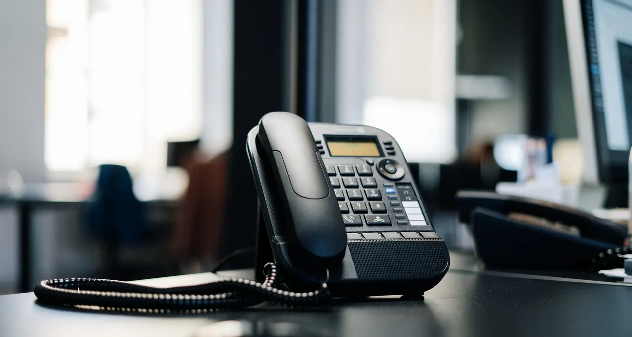 A black IP desk phone on an office desk, with a second handset and monitors soft in the background.