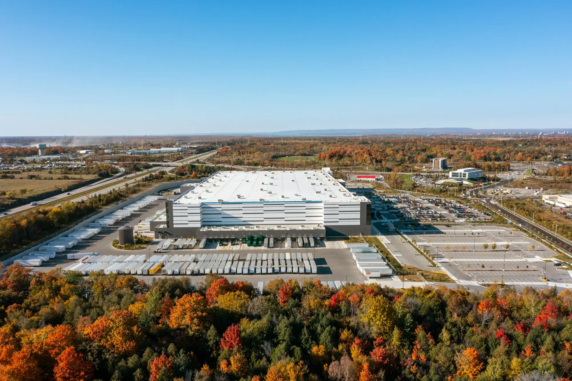 A large distribution center from the air, with long rows of trailers at the docks and in the yard and an autumn forest in front.