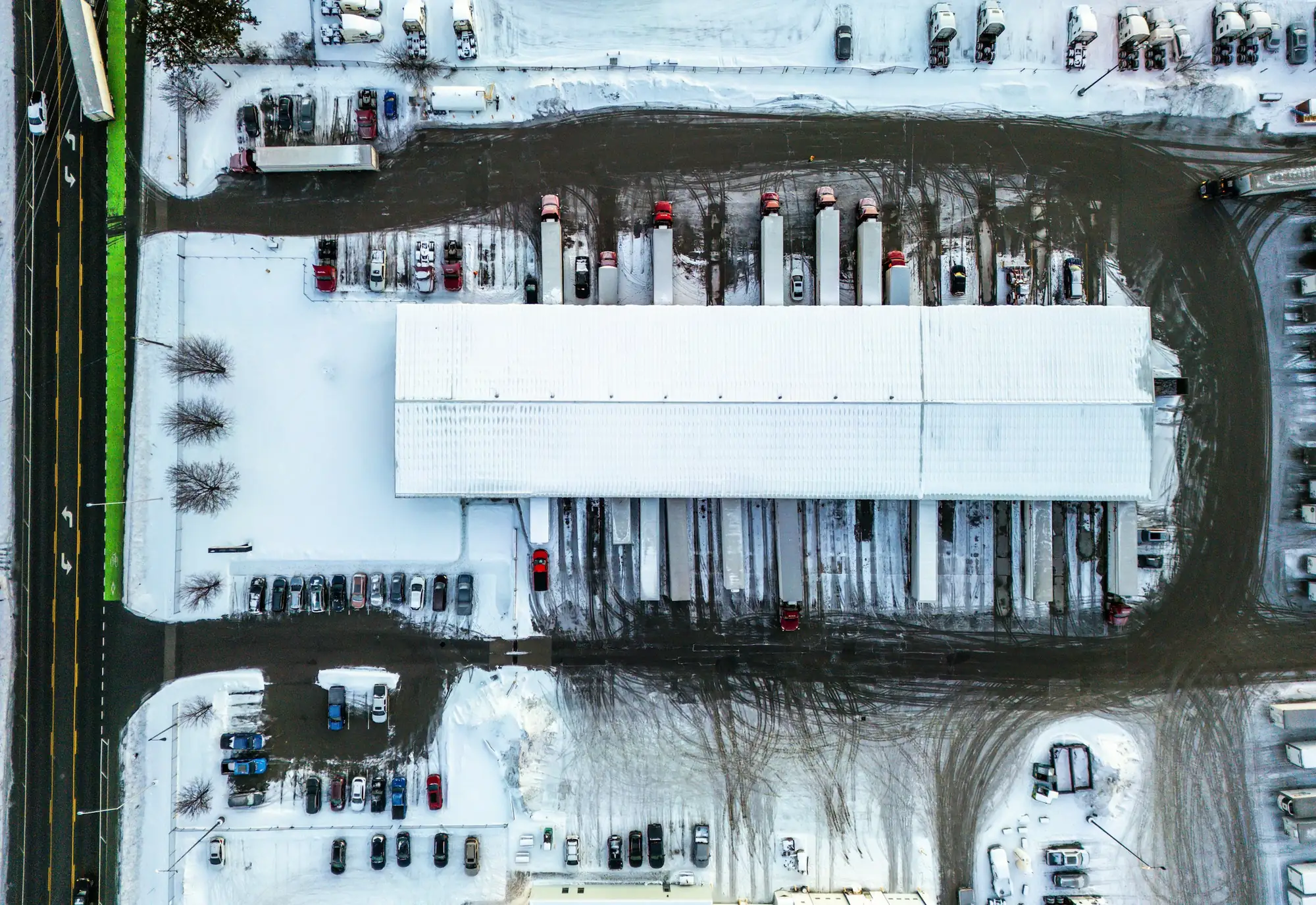 A large distribution center seen from directly above in winter, with trailers backed into every dock door and cars in the lots around it.
