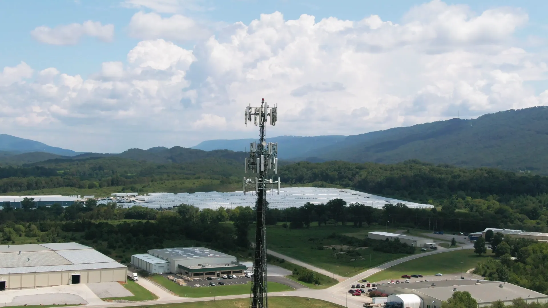 A cell site with rows of carrier antennas stands beside a business park of large warehouses and offices, with wooded hills behind it, on a bright day.