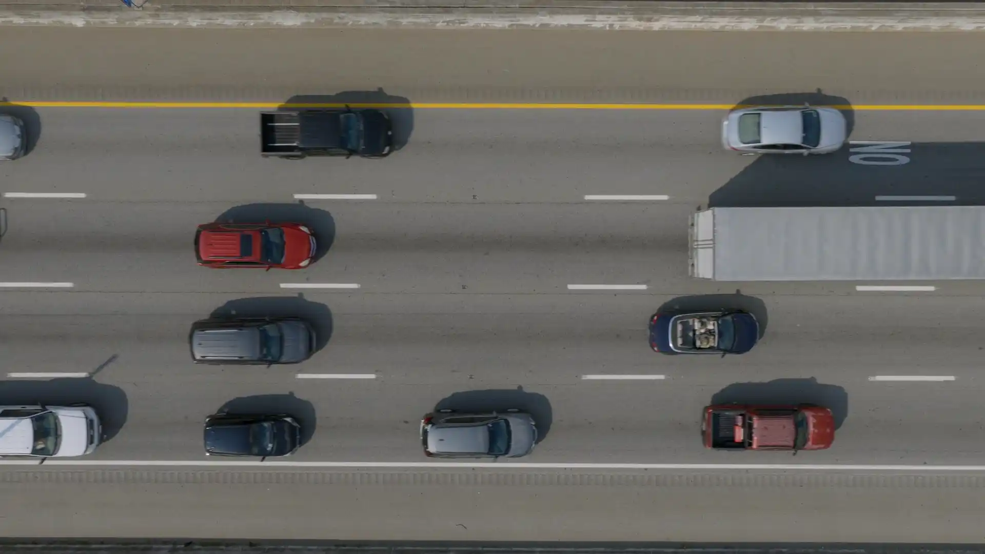 An interstate seen from directly above, with a tractor-trailer and cars moving through the lanes in bright daylight.