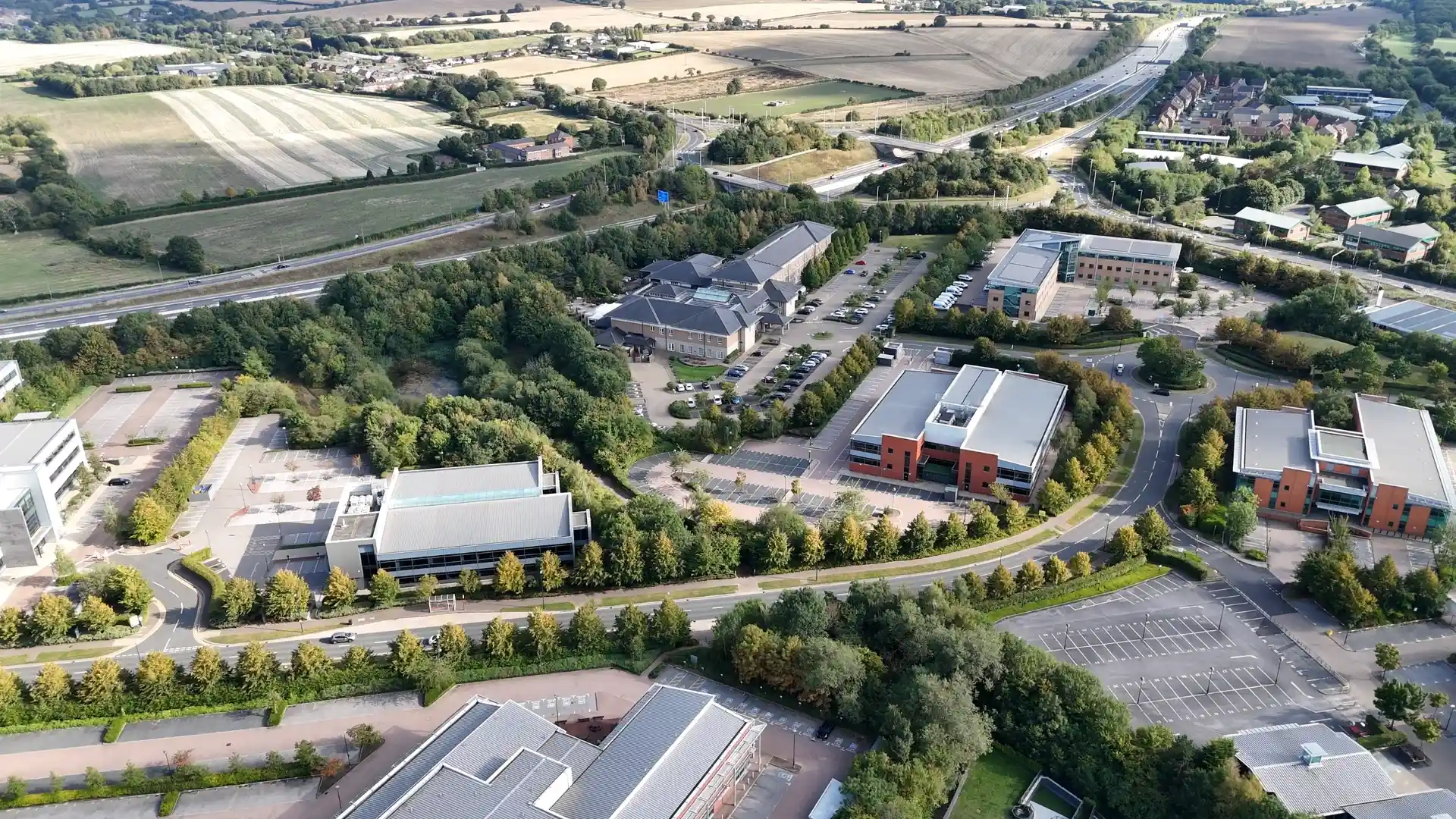 A drone view of a business campus: office buildings, parking lots and tree-lined roads on a sunny afternoon.
